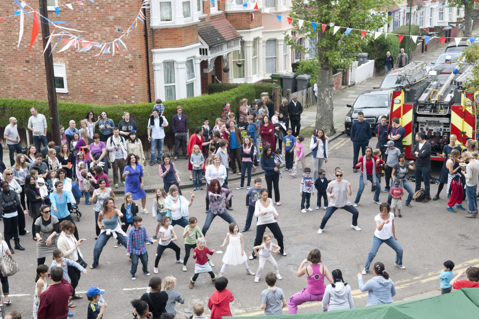 A large group of people of all ages participating in a street dance event on a residential street, with a fire truck parked nearby