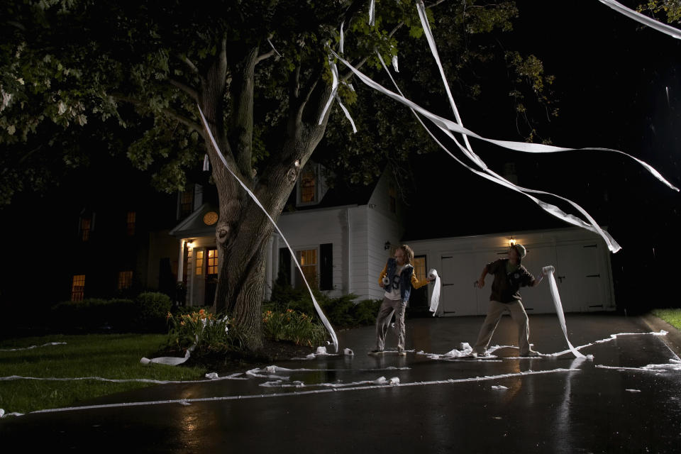 Two people decorate a tree with toilet paper at night in front of a house, surrounded by scattered paper