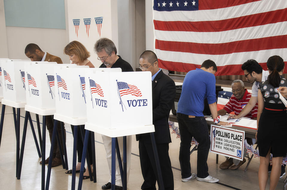 People voting in privacy booths at a polling place, with election staff assisting and an American flag in the background